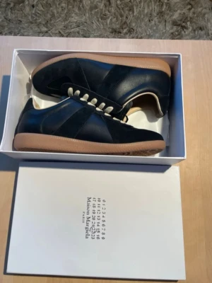 Maison Margiela svarta sneakers - Maison Margiela sneakers i svart läder och mocka med beige innersula och vita snören. Klassisk låg modell med gummisula i brunt. Snygg och stilren design som passar till det mesta. Levereras i originalkartong.