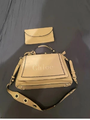 Unik Beige handväska från Chloé - Snygg unik beige handväska från Chloé med dekorativt broderad kant och präglad logga framtill. Väskan har både handtag och bred axelrem med stora guldfärgade öljetter. Tillverkad i skinn och har en klassisk, fyrkantig form. Väskan utan defeker alls plånboken lite se bild. Använd gärna köp nu❤️
