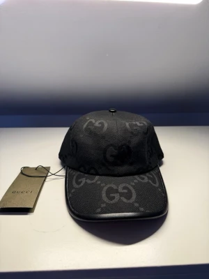 Svart keps från Gucci med GG-mönster - Snygg svart keps från Gucci med klassiskt GG-monogram över hela kepsen. Kepsen har böjd skärm och är tillverkad i ett slitstarkt tygmaterial med detaljer i läder. Perfekt accessoar för en trendig streetstyle-look.