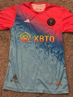 Inter Miami matchtröja Messi #10 - Säljer en Inter Miami matchtröja från Adidas med Messi #10 på ryggen. Tröjan har en unik färgkombination med rosa axlar och ärmar samt blått och turkost mönster på framsidan. Tryckta loggor och sponsor på bröstet. Perfekt för fotbollsfans!