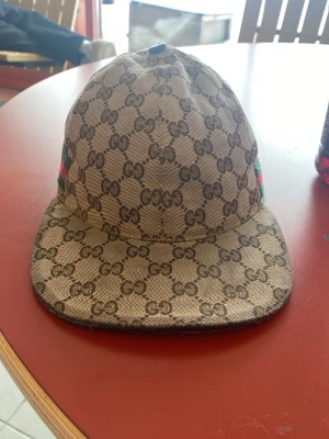 Gucci keps  - Säljer en beige keps från Gucci med klassiskt GG-monogram över hela kepsen. Kepsen har böjd skärm och detaljer med grönt och rött band på sidan. Materialet är canvas och kepsen har justerbar rem baktill.
