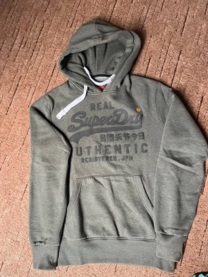 Grå hoodie från Superdry  - Säljer en grå hoodie från Superdry med stort tryck på bröstet och japanska tecken. Hoodien har en klassisk känguruficka, ribbade muddar och vit dragsko i huvan. Mjuk insida i fleece och avslappnad passform. Perfekt för chill dagar eller streetwear-stil.