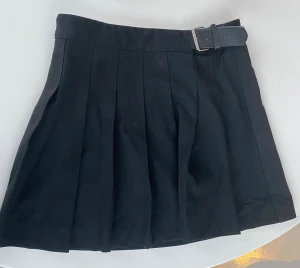 Svart plisserad kortkjol med bälte - Snygg svart kortkjol med plisserade veck och ett inbyggt bälte i midjan. Kjolen har en klassisk look och passar perfekt till många olika stilar. Tillverkad i ett mjukt tyg som ger en bekväm känsla.