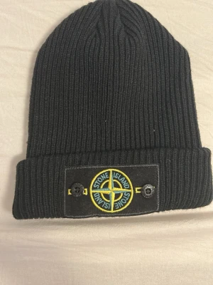 Svart ribbstickad mössa Stone Island - Säljer en svart ribbstickad mössa från Stone Island med uppvikt kant och den klassiska logotypen framtill i gult och grönt på svart patch. Mössan har en clean och stilren look, perfekt för dig som gillar streetwear och vill ha något som sticker ut.