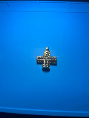 Guldfärgat korshalsband med stenar - Snyggt halsbandshänge i form av ett kors, guldfärgat med glittrande stenar infattade på framsidan. Perfekt för dig som gillar detaljer och vill ha något som sticker ut. Hänget har en klassisk och stilren design.