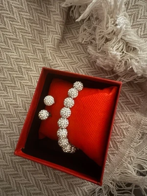 Glittrigt armband med matchande örhängen - Säljer ett silvrigt armband med stora, runda glittrande stenar och matchande örhängen. Armbandet har en klassisk design med flera gnistrande stenar på rad. Perfekt för dig som gillar att sticka ut med accessoarer som verkligen syns.