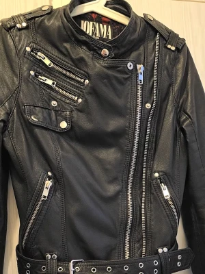 Svart biker skinnjacka Jofama - Snygg svart skinnjacka från Jofama i klassisk bikerstil med flera dragkedjor och bälte i midjan. Jackan har ståkrage med tryckknapp och coola detaljer i silverfärgade metalldelar. 