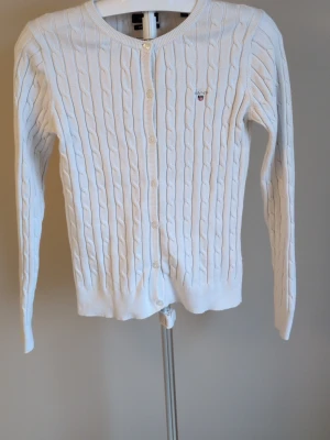 Vit kabelstickad kofta från Gant - Kabelstickad vit kofta från Gant med rund halsringning och knäppning framtill. Liten broderad logga på bröstet. Långa ärmar och ribbade muddar vid ärmslut och nederkant. Perfekt för lager på lager och klassisk stil.