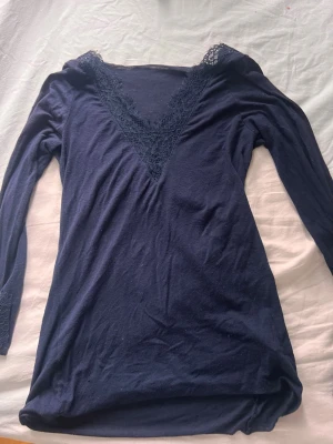 Mörkblå långärmad topp från intimissimi med spets - Mörkblå långärmad topp med djup v-ringning och dekorativ spets runt halsringningen och ärmsluten. Sitter tight och har en feminin, elegant look som passar perfekt till jeans eller kjol.