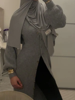 Lång grå stickad kofta - Snygg lång grå stickad kofta med ribbad struktur och knappar hela vägen framtill. Koftan har ballongärmar och sitter figurnära upptill. Perfekt för lager-på-lager och enkel att styla med olika outfits.