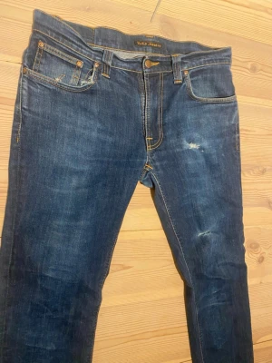 Blå jeans från Nudie Jeans - Säljer ett par klassiska blå jeans från Nudie Jeans med snygga slitningar och kontrastsömmar. Modell thin fin och Modellen har normal passform och raka ben, med Nudie Jeans ikoniska broderi på bakfickorna. Tillverkade i slitstarkt jeanstyg av bomull. Tveka inte att fråga eller ge ditt pris!