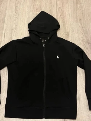 Svart hoodie från Ralph Lauren - Svart hoodie med dragkedja från Ralph Lauren. Klassisk design med huva, två fickor framtill och den ikoniska vita Polo-loggan broderad på bröstet. Perfekt för en avslappnad och stilren look.