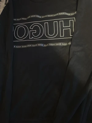 Svart sweatshirt från Hugo - Säljer en svart sweatshirt från Hugo med stort vitt logotryck på bröstet och små loggor runtom. Tröjan har rund halsringning och långa ärmar. Perfekt för dig som gillar stilrena och trendiga plagg med tydlig branding.