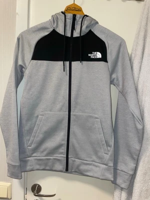Grå hoodie från The North Face - Snygg grå hoodie med svart panel upptill från The North Face. Dragkedja hela vägen, två fickor fram och justerbar huva med snören. Perfekt för chill eller när du vill ha en clean streetstyle-look. Tröjan är helt ny med prislapp
