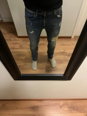 Blå slim fit jeans med patchar - Säljer ett par blå slim fit jeans med slitningar och coola patchar på låret. Jeansen har klassisk femficksdesign och detaljerade sömmar. Perfekta för dig som gillar en trendig och avslappnad streetstil.
