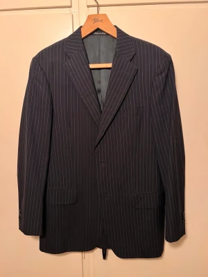 Rose & Born suit jacket - Snygg mörkblå kavaj från exklusiva Rose & Born, tillverkad i Italien. Kavajen har smala vita kritstrecksränder, klassisk passform med två framfickor med lock och bröstficka. Perfekt för en dressad look och tillverkad i ett tunt, elegant tyg. Storlek 48. Nypris: 14 000kr