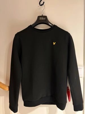 Svart sweatshirt från Lyle & Scott - Klassisk svart sweatshirt från Lyle & Scott med rund halsringning och den ikoniska gula örnloggan broderad på bröstet. Tröjan har långa ärmar och ribbade muddar vid ärmslut och nederkant. Passar en person perfekt mellan 160-175 