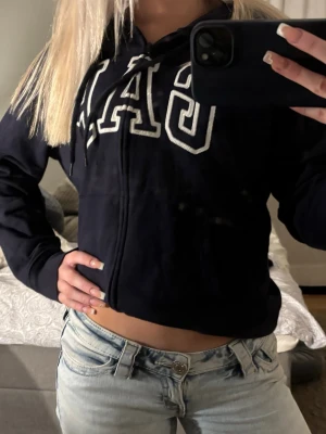 Mörkblå hoodie från GAP med dragkedja - Mörkblå hoodie från GAP med vit broderad logga på bröstet. Hoodien har dragkedja framtill, snörning i huvan och en croppad passform. Perfekt för en chill och avslappnad stil.