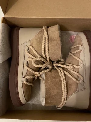 Beige moon boots från Inuikii - Säljer ett par snygga moon boots från Inuikii i beige mocka och läder med grov brun sula. Skorna har rejäla snören och en cool, chunky design som passar perfekt till vintern. Fodrade för extra värme och stilpoäng.
