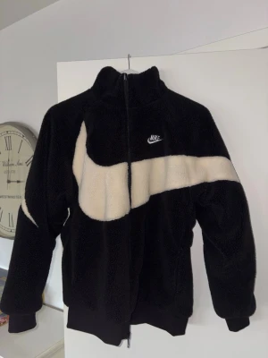 Nike Reversible Swoosh Fleece Jacka/Kofta - Säljer en svart  Reversible swoosh fleece jacka/kofta från Nike med stor vit Swoosh-logga som går över både fram- och baksidan. Jackan är även ”Reversible” (Dvs man kan även vända den ut och in och ta på sig den.) med en dragkedja som kan snurras mot andra hållet när man vänder ut och in på jackan. Jackan har hög krage, dragkedja och muddar vid ärmslut och nederkant. Perfekt för dig som vill ha en sportig och trendig look.