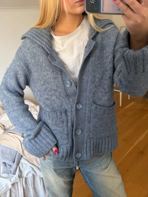 Blå stickad kofta med knappar - Mysig blå stickad kofta med stora knappar framtill och två fickor. Koftan har en avslappnad passform, ribbade muddar och en bred krage. Perfekt att slänga över en t-shirt för en chill look. Stor i storleken!