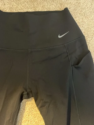 Svarta leggings från Nike - Säljer ett par svarta leggings från Nike med hög midja och diskret Nike-logga på vänster sida. Tillverkade i stretchigt material som sitter skönt och har fickor på sidorna. Perfekta för träning eller chill dagar.