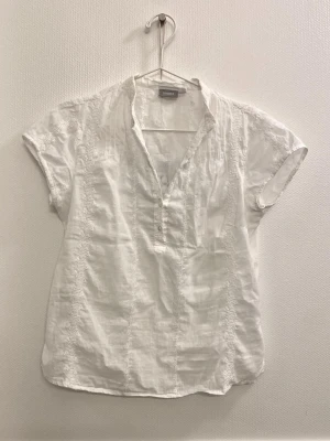Vit blus med knappar från Lindex - Vit blus från Lindex med korta ärmar och v-ringning. Blusen har dekorativa sömmar framtill och knäppning med två knappar. Tillverkad i ett lätt och luftigt tyg.