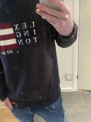 Mörkblå stickad tröja Lexington - Säljer en mörkblå stickad tröja från Lexington med vit och röd detalj samt text framtill. Tröjan har ribbstickade muddar vid ärmslut och nederkant. Perfekt för dig som gillar klassisk och stilren design.