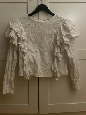 Vit blus med volang och broderi - Superfin vit blus med broderade detaljer och volanger längs axlarna. Blusen har långa ärmar och dekorativa små bollar på volangkanterna. Använt fåtal gånger och säljer pga att den är lite liten för mig🤍🤍