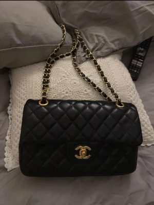 Chanel svart quiltad handväska - Säljer en klassisk svart quiltad handväska  med guldfärgade detaljer och kedjerem. Väskan har dubbla lock, ikonisk CC-logga framtill och insida i vinrött läder. Perfekt för dig som vill ha en tidlös och lyxig accessoar.