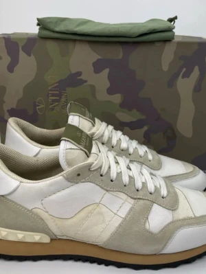 Valentino Garavani vita sneakers - Snygga vita sneakers från Valentino Garavani med beige och ljusgrå detaljer. Skorna har en klassisk låg silhuett, mocka- och läderpaneler samt kamouflagemönster. Gummisula med nitar på hälen och vita skosnören. Perfekta för dig som gillar exklusiv streetstyle.