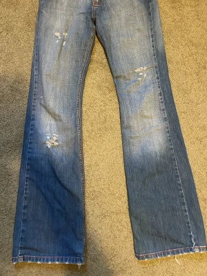 Blå bootcut jeans med slitningar - Säljer ett par blå bootcut jeans med slitningar på framsidan och detaljerade bakfickor. Jeansen har en klassisk femficksdesign och är gjorda i denim med lätt tvättad. Inerbenslängd: 85 midja tvärs över: 35 