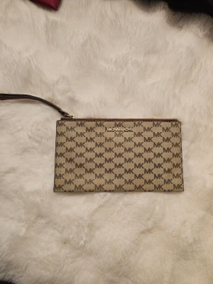 Beige kuvertväska Michael Kors - Snygg beige kuvertväska från Michael Kors med klassiskt MK-mönster över hela väskan. Väskan har guldfärgad dragkedja och logga framtill samt en avtagbar handledsrem i brunt läder. Insidan är fodrad med ljust tyg och har en liten ficka.