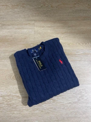 Mörkblå kabelstickad tröja Polo Ralph Lauren - Mörkblå kabelstickad tröja från Polo Ralph Lauren med rund halsringning och klassisk röd broderad logga på bröstet. Tröjan har ribbade muddar och är tillverkad i mjukt material som ger en stilren look.
