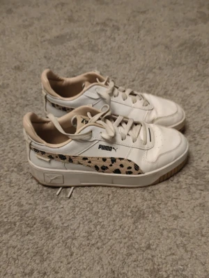 Puma vita sneakers med leoparddetalj - Snygga vita sneakers från Puma med beige och svarta leopardmönstrade detaljer på sidan. Skorna har klassisk låg modell, vita snören och beige insida. Tillverkade i läder och syntetmaterial med gummisula. Perfekta för en trendig streetstyle-look.