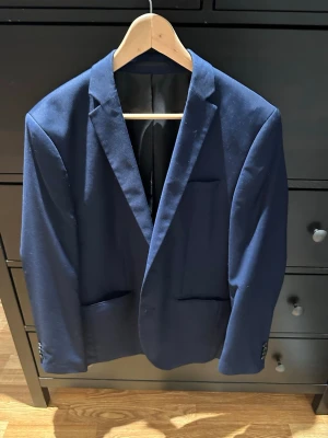 Mörkblå klassisk kavaj Jack&Jones - Snygg mörkblå kavaj med smal passform och klassiska slag. Kavajen har två framfickor och knappar vid ärmsluten. Perfekt för en stilren look. Materialet känns slätt och bekvämt.