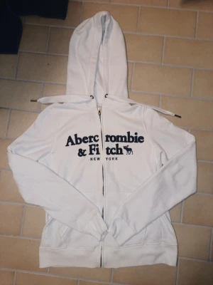 Vit hoodie/zip up från Abercrombie & Fitch  - Vit zip-hoodie från Abercrombie & Fitch med stor svart logga och text på bröstet. Klassisk huva med dragsko och fickor framtill. Perfekt för en chill och avslappnad stil.