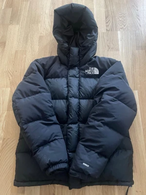 Svart dunjacka från The North Face - Svart dunjacka från The North Face med huva och broderad logga på bröstet och ryggen. Jackan är quiltad och har en pufferstil med 650 fill power, perfekt för kalla dagar. Klassisk och stilren design som funkar till det mesta.
