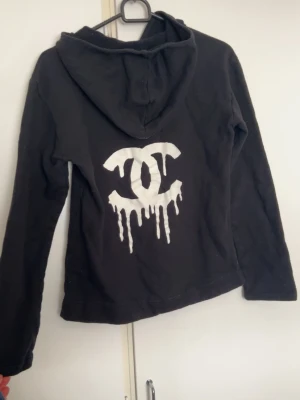 Svart hoodie med drip-logga - Svart hoodie med stor vit drip-inspirerad logga på ryggen och klassisk känguruficka framtill. Hoodien har huva och långa ärmar, perfekt för en chill och streetig look. . Passar dig som gillar edgy och unik stil. Passar XS/S