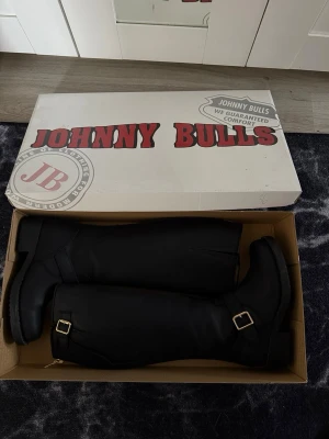 Svarta stövlar från Johnny Bulls - Oanvända, Snygga svarta stövlar från Johnny Bulls med guldfärgade spännen vid ankeln. Stövlarna är höga, har en klassisk form och är tillverkade i slätt läder. Perfekta för dig som vill ha en stilren och tidlös look.