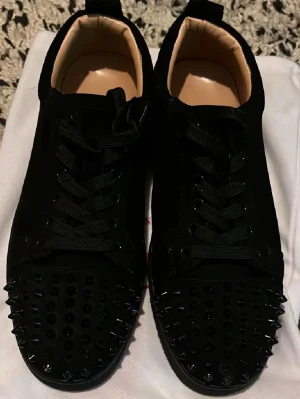 Svarta spikes-sneakers Christian Louboutin - Svarta sneakers i mocka från Christian Louboutin med svarta nitar och spikes över hela tån. Klassisk låg modell med svarta skosnören och beige innersula med guldfärgad logga. Riktigt edgy och sticker ut i mängden.