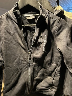 Svart fleecejacka från Sail Racing - Svart fleecejacka från Sail Racing med dragkedja framtill och hög krage. Jackan har logga broderad på bröstet och två sidofickor med dragkedja. Perfekt för lager-på-lager och riktigt skön att ha på sig.