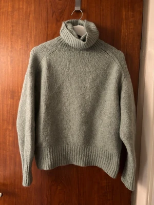 Grön stickad polotröja - Mysig grön stickad polotröja med hög krage och långa ärmar från Zara. Tröjan har en avslappnad passform och breda ribbstickade muddar vid ärmslut och nederkant. Perfekt för kalla dagar och enkel att matcha med jeans eller kjol.