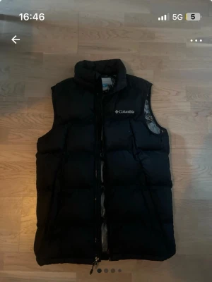 Svart dunväst från Columbia - Snygg svart dunväst från Columbia med hög krage och dragkedja framtill. Västen har ett puffigt quiltat mönster och är fodrad med glansigt material på insidan. Perfekt för lager-på-lager och håller dig varm under kyliga dagar.