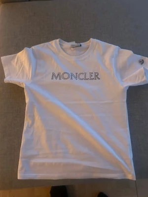 Vit Moncler t-shirt med logga - Snygg vit t-shirt från Moncler med klassisk logga tryckt på bröstet och Moncler-märke på ärmen. T-shirten har rund hals och korta ärmar, perfekt till en clean och stilren look. Tillverkad i mjuk bomull för maximal komfort. Skall tilläggas att jag inte använder den eftersom den är för liten för mig. Priset kan diskuteras. Större chans att de sänks vid en snabb affär.