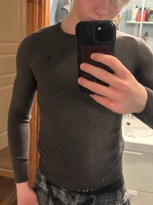 Mörkbrun stickad tröja Morris - Tröjan tvättas fel och är numera som en storlek xs-xxs !!! Mörkgrå stickad tröja från Lexington med rund halsringning och diskret broderad logga på bröstet. Tröjan har långa ärmar och ribbade muddar vid ärmslut och nederkant. Perfekt för lager-på-lager under kyliga dagar.