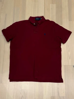 Vinröd pikétröja från Polo Ralph Lauren - Klassisk vinröd pikétröja från Polo Ralph Lauren med krage, korta ärmar och knappslå framtill. Tröjan har den ikoniska lilla logotypen broderad på bröstet. Tillverkad i bomull och har en normal passform.