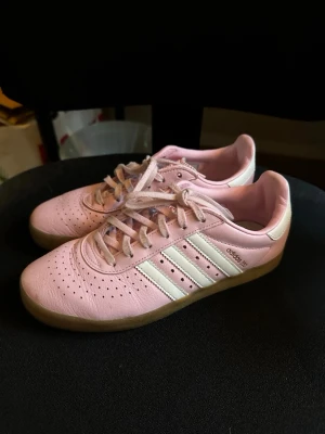 Adidas 350 rosa sneakers med snörning - Säljer ett par Adidas 350 sneakers i ljusrosa skinn med klassiska vita ränder på sidorna och beige gummisula. Skorna har perforerat mönster på tån och snörning framtill. Perfekt för dig som gillar sportig och clean stil.