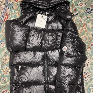 Svart Moncler pufferjacka med huva storlek m och L  - Svart pufferjacka från Moncler med glansig finish, huva och dragkedja framtill. Jackan har Moncler-logga på ärmen, praktiska fickor med dragkedja och tryckknapp, samt quiltad design. Perfekt för kalla dagar och riktigt snygg streetstil.   
