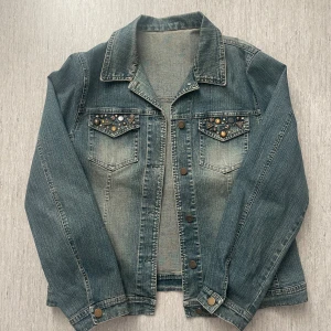Blå jeansjacka med snygga detaljer - Snygg blå jeansjacka med nitar och metallknappar på bröstfickan. Jackan har långa ärmar, knappstängning framtill och sitter ganska baggy. 